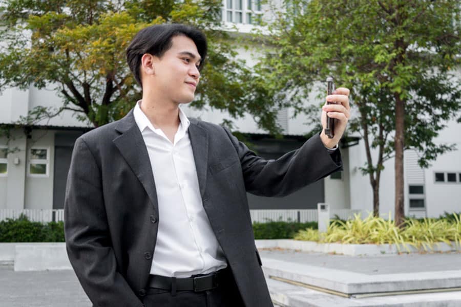 A man in a dark suit stands outdoors, smiling and holding up a smartphone to take a selfie for video verification, with trees and a modern building in the background—perfectly capturing the process many local businesses follow when setting up their Google Business Profile.