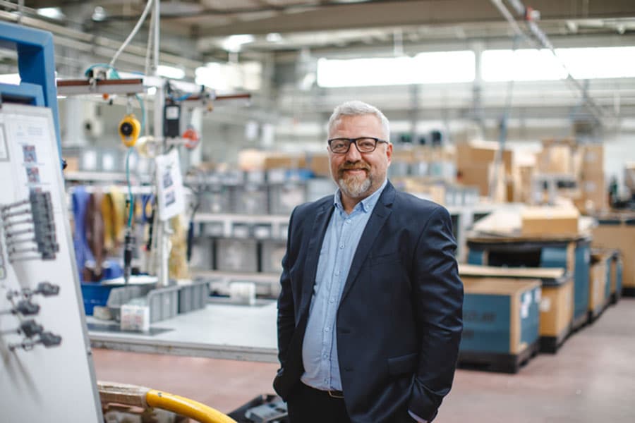 A middle-aged man with gray hair and a beard, wearing glasses and a suit, stands smiling in a factory workspace—an ideal scene for branding a business by creating a memorable brand amid industrial equipment, boxes, and machinery.