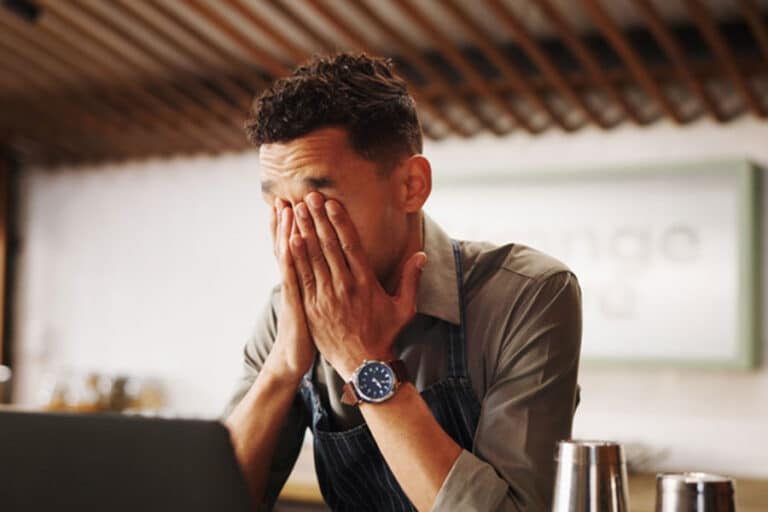 A man sits at a table with his hands covering his face, appearing stressed or overwhelmed—perhaps the result of dealing with bad clients. A laptop and some metal containers are in front of him, hinting that client sorting might be an ongoing challenge.