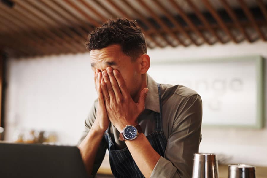 A man sits at a table with his hands covering his face, appearing stressed or overwhelmed—perhaps the result of dealing with bad clients. A laptop and some metal containers are in front of him, hinting that client sorting might be an ongoing challenge.