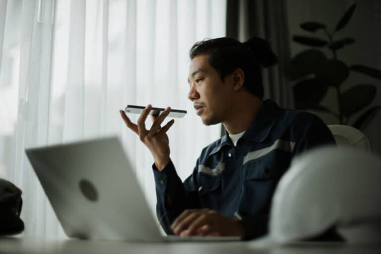 A man sits at a desk with a laptop, holding a smartphone near his mouth as if using voice commands for AI search optimization. A white hard hat rests on the desk, and light filters through sheer curtains behind him.