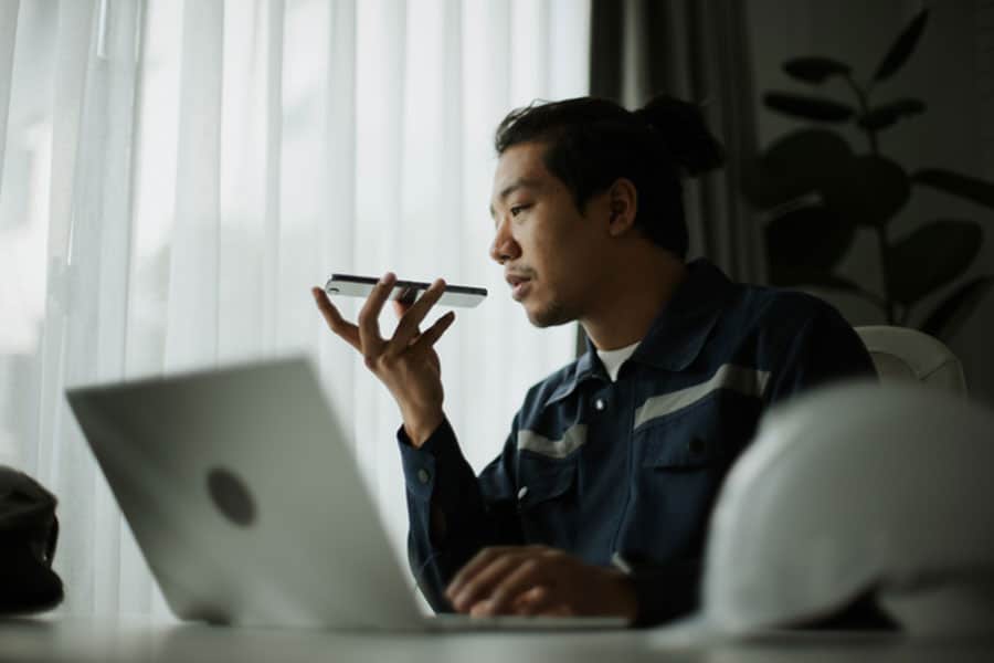 A man sits at a desk with a laptop, holding a smartphone near his mouth as if using voice commands for AI search optimization. A white hard hat rests on the desk, and light filters through sheer curtains behind him.