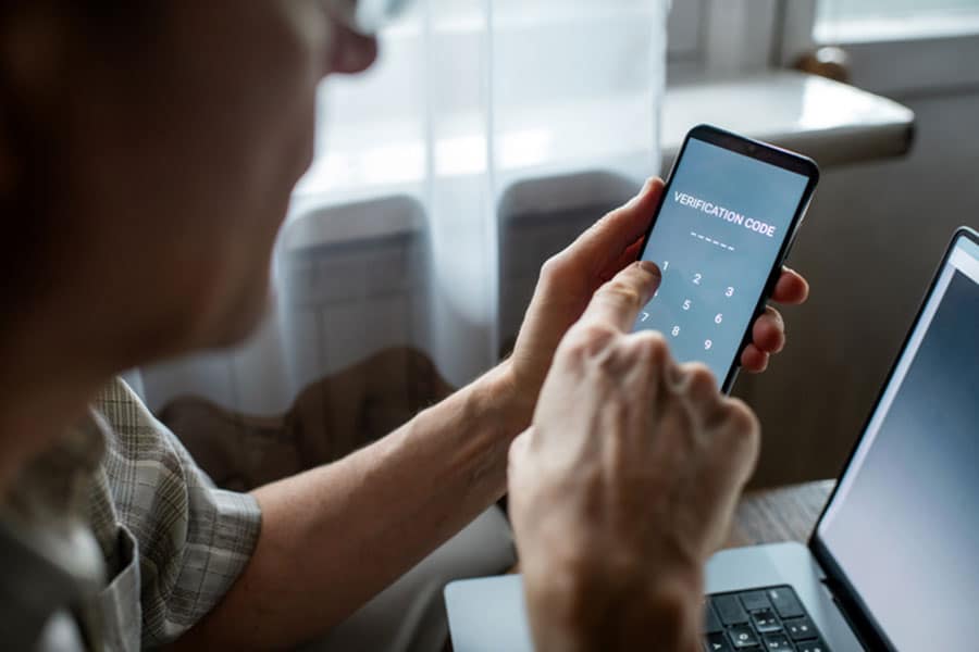 A person enters a verification code on a smartphone, sitting beside an open laptop with light streaming through a window in the background—perhaps completing video verification for their Google Business Profile to support local businesses.