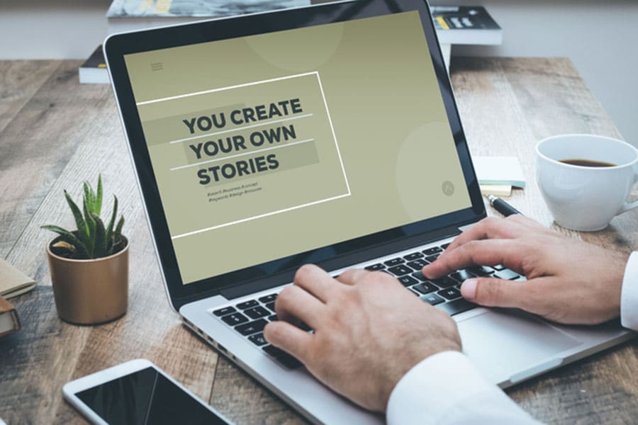 A person types on a laptop at a wooden desk, crafting their latest story. The screen displays the text YOU CREATE YOUR OWN STORIES. A notebook, pen, smartphone, and cup of coffee are on the desk—essentials for anyone passionate about blogging and creative expression.