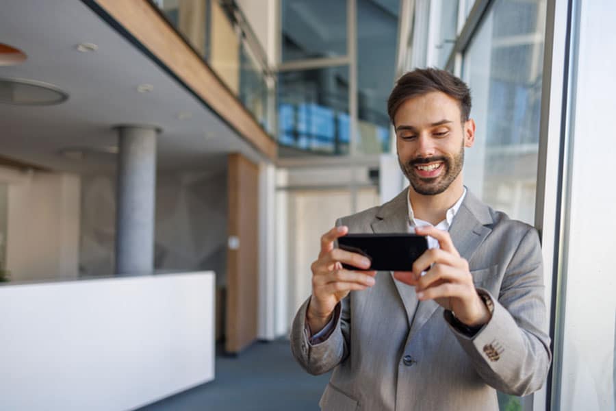 A man in a light gray suit stands indoors by large windows, smiling while using a smartphone. He appears to be assisting local businesses with Google Business Profile video verification. A modern office lobby with glass walls and natural light is visible in the background.