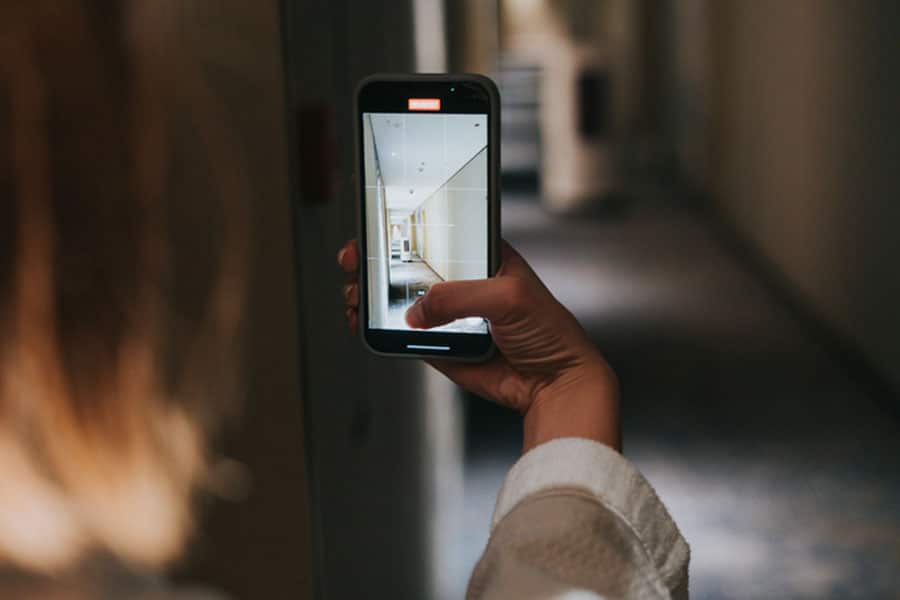 A person holds a smartphone and takes a photo of a hallway with beige walls and lights. The phone screen displays the hallway, creating a mirrored effect, similar to the process used in Google Business Profile Video Verification for local businesses.